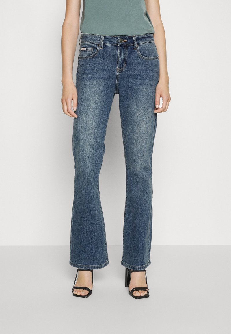 Žena v bledě modrých bootcut džínách a černých páskových lodičkách na vysokém podpatku, stojící na neutrálním světlém pozadí.