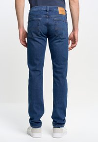 Mees, kes kannab siniseid sirge säärega teksapükse ja valgeid tossusid, nähtud tagant tavalise valge taustaga.