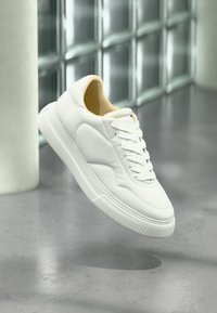 Άσπρο sneaker χαμηλού προφίλ με κορδόνια, αιωρούμενο πάνω από γκρι πάτωμα, με γυάλινο τοίχο από τουβλάκια και λευκή κυλινδρική κολόνα στο φόντο.