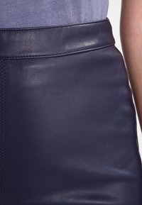 Marint blå läderkjol med en slät yta, har hög midja och synlig sömnad längs sömmen. Minimalistisk design utan ytterligare detaljer.