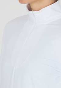 Weißes Sportoberteil mit hohem Kragen, Viertel-Reißverschluss und glattem, dehnbarem Material. Mit feinen Nahtdetails.