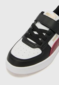 Sneaker en cuir noir et blanc avec un bout perforé, une sangle texturée noire et un accent rouge contrastant. Dispose de lacets noirs plats et d'un logo.
