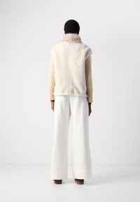 Persona che indossa un gilet con collo in pelliccia color crema, un top a maniche lunghe beige, pantaloni bianchi a gamba larga e scarpe marroni, in piedi contro uno sfondo bianco.