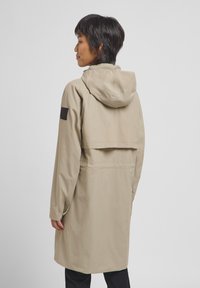 Beige waterdichte trenchcoat met een capuchon, voorzien van een rugflap, lange mouwen en een logo-opdruk op de linkerarm. Glad, textuurachtig fabric.