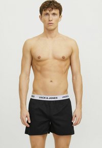Shorts noirs avec une ceinture élastique blanche arborant "JACK & JONES" en lettres noires en gras. Tissu lisse, coupe ample, longueur mi-cuisse.