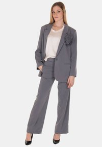 Completo da donna grigio con blazer sartoriale caratterizzato da un dettaglio di fiore in tessuto, abbinato a pantaloni a gamba larga. Sotto, una camicetta di colore crema.