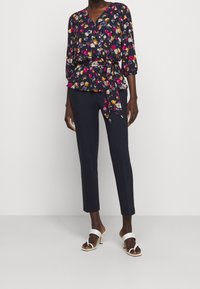 Peplumtopp med blommigt mönster, djup V-ringning, trekvartsärmar och knyt i midjan, kombinerad med figursydda mörka byxor och vita högklackade sandaler.