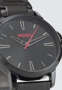 Montre en acier inoxydable noir avec un cadran noir mat, des index horaires argentés et un logo "HUGO" rouge. Dispose d'une aiguille des secondes rouge et d'un bracelet à maillons multiples.