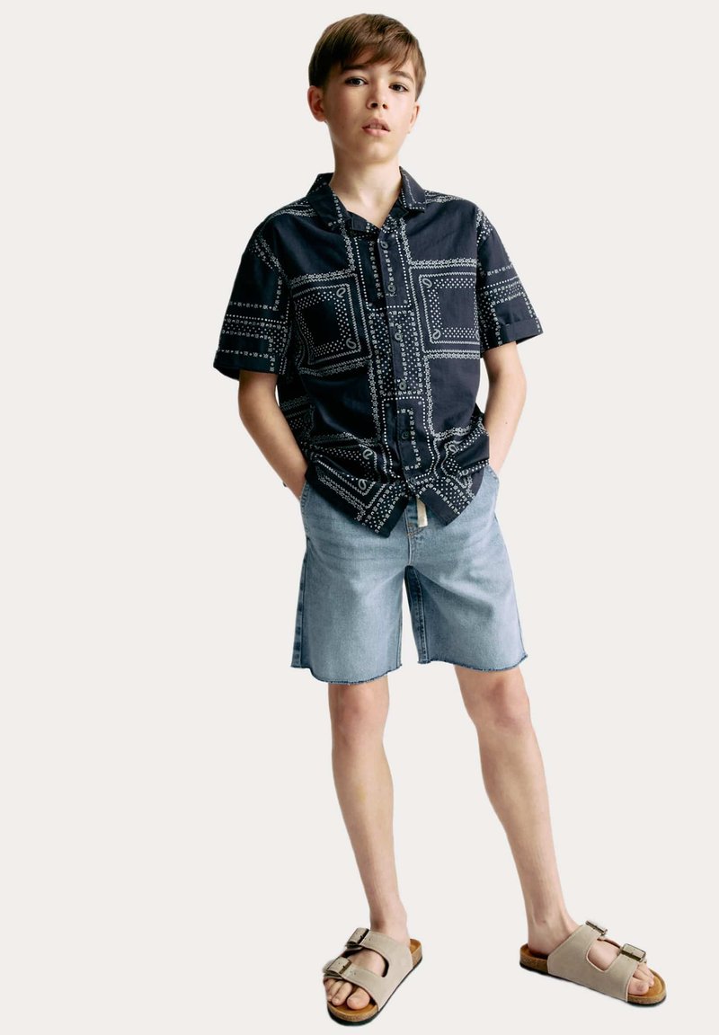 Camisa negra de manga corta con acentos de patrones blancos, pantalones cortos de mezclilla azul claro y sandalias beige con detalle de hebilla.
