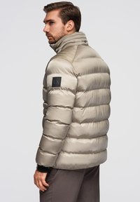 Beige puffjakke med høj krave, quiltet design og et sort logo patch på venstre ærme. Lavet af et blankt, glat materiale.
