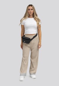 Zwarte heupband met logo, crossbody gedragen, gecombineerd met beige geribde wijde broek en een witte aansluitende t-shirt. Witte sneakers zichtbaar.