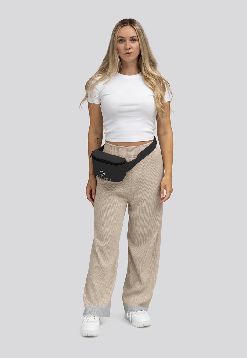 Zwarte heupband met logo, crossbody gedragen, gecombineerd met beige geribde wijde broek en een witte aansluitende t-shirt. Witte sneakers zichtbaar.