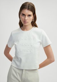 Bílý bavlněný tričko s krátkým rukávem s texturovaným vyšitým nápisem "BIBLIO-NS THEQUE -ULRICO" na přední straně. Krátký střih.