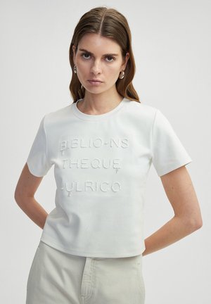 T-shirt blanc en coton à manches courtes avec un texte brodé texturé indiquant "BIBLIO-NS THEQUE -ULRICO" sur le devant. Design court.