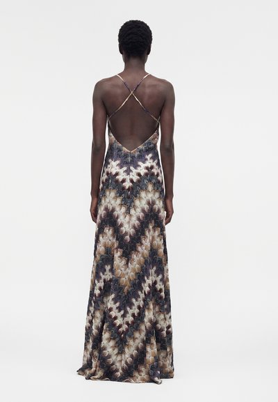 Γυναίκα που φοράει ένα αμάνικο, μακρύ φόρεμα με λεπτές λουρίδες που διασταυρώνονται στην πλάτη και πολύχρωμο ζιγκ-ζαγκ μοτίβο σε αποχρώσεις καφέ, μαύρου και μπεζ.