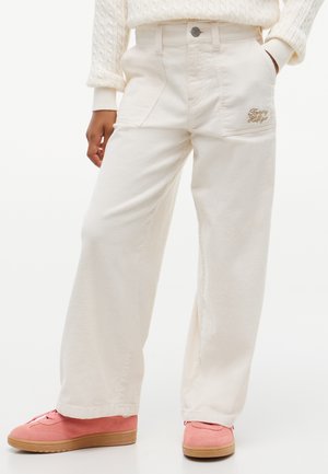 Personne portant un pantalon crème à jambes larges avec poches avant et un pull, associé à des baskets roses en daim avec semelles en gomme.