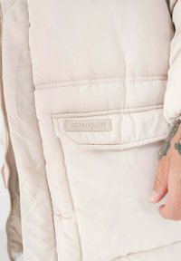 Lichtbeige pufferjack met een gequilt ontwerp, voorzien van een zijzak en een gegraveerd "SIXTH JUNE" label. Glad, iets glanzend stof.