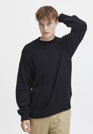 Black knitted sweater with a crew neckline, loose fit, and ribbed cuffs. Soft texture and casual silhouette. Worn with beige pants.