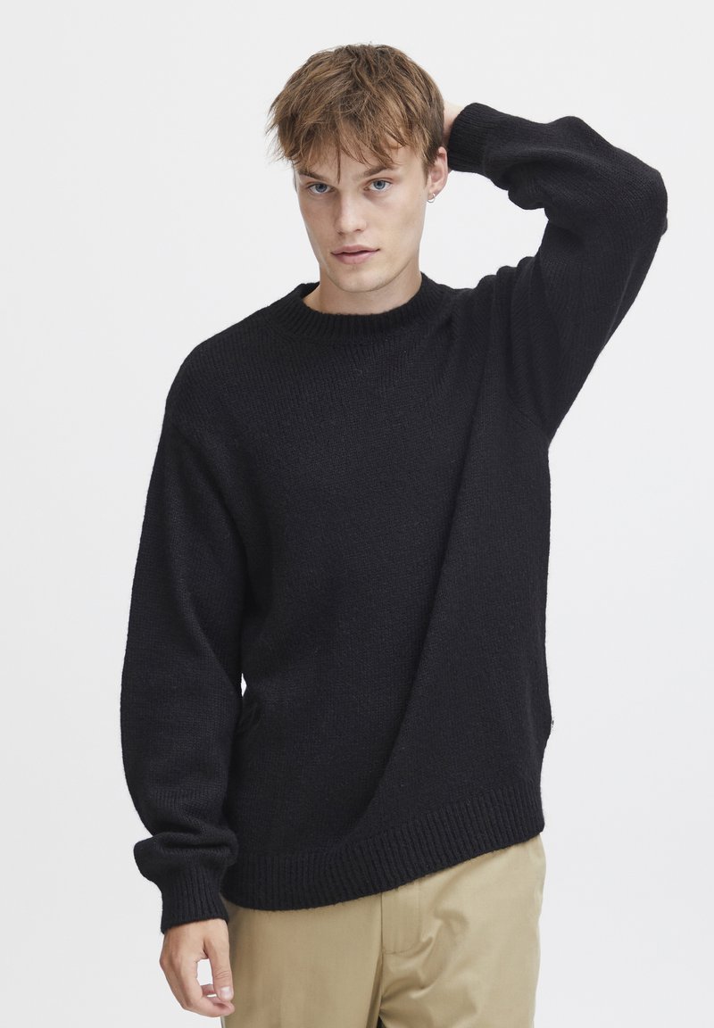 Suéter negro de punto con cuello redondo, corte holgado y puños acanalados. Textura suave y silueta casual. Combinado con pantalones beige.