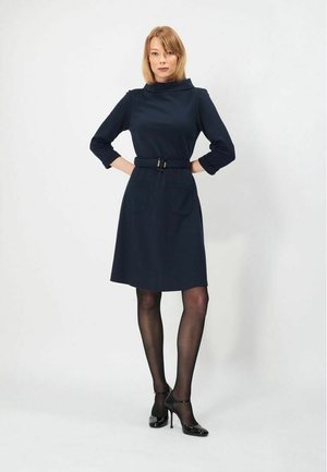 Kvinde står selvsikkert iført en knælang mørkeblå kjole med bælte, sorte gennemsigtige strømpebukser og sorte højhælede sko mod en ensfarvet baggrund.