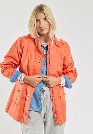Veste imperméable orange avec capuche, poches avant et poignets ajustables, superposée sur une chemise en denim et un haut à rayures à manches longues.