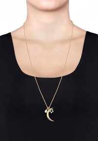 Collier en pendentif doré présentant un design de croissant de lune et d'étoile, orné d'une petite pierre précieuse, exposé sur un haut noir contre un fond clair.
