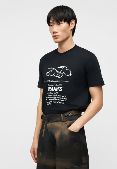 Άνδρας με μαύρο μπλουζάκι με σχέδιο Snoopy και κείμενο, συνδυασμένο με μαύρο και καφέ παντελόνι cargo με βάψιμο tie-dye, στέκεται μπροστά σε απλό φόντο.