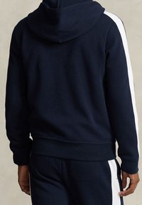 Marinblå hoodie tillverkad av textil med struktur, med vita sidosträck och ribbad mudd och nederkant. Inkluderar känguruficka.
