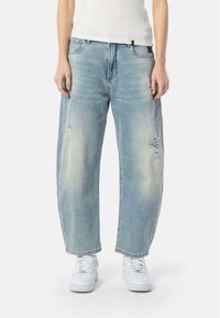 Jean en denim bleu clair avec une coupe ample, des détails usés et un design classique à cinq poches, associés à des baskets blanches.
