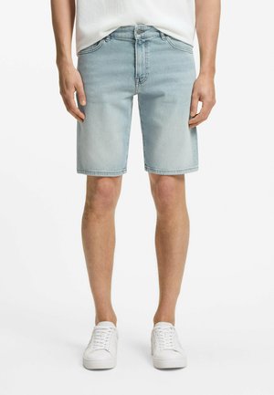 Mann trägt hellblaue Jeansshorts und weiße Sneaker, steht vor einem einfarbigen weißen Hintergrund.