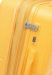 Gelbe Hartschalenreisetasche mit strukturierten Oberfläche, zwei silbernen Reißverschlüssen, integriertem Zahlenschloss und Markierungsdetails an den Reißverschlüssen.