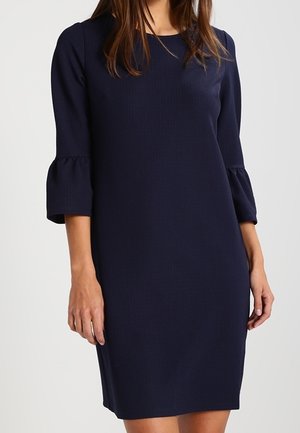 Femme portant une robe bleu marine mi-longue aux manches évasées trois-quarts et au col rond, debout devant un fond uni.