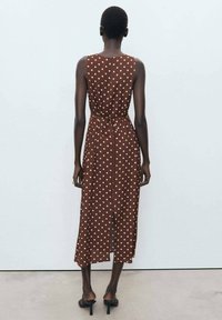 Brown polka dot dress with a fitted waist, sleeveless design, mid-calf length, and side slit; fabric appears smooth and lightweight.