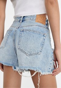 Shorts de denim azul claro desgastados con dobladillo deshilachado, que cuentan con dos bolsillos traseros y un parche de cuero marrón con el nombre de la marca en la cinturilla.