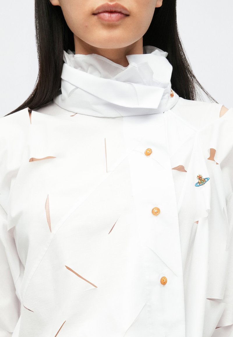 Vit skjorta med lager-på-lager utskurna detaljer, kompletterad med orange knappar och en liten broderad design. Krusad krage som tillför textur.