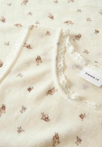 Blommönstrad krämfärgad linne med spetsdetaljer, tillverkat av mjukt material. Har små, dämpade blommor och en etikett med texten "name it."