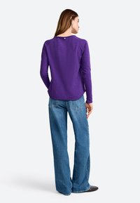 Purple long-sleeve shirt with a rounded neckline, featuring a small button detail at the back. Paired with light blue wide-leg jeans.