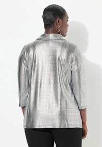 Silberner, schlangenförmiger Oberteil mit Kragen, Dreiviertelärmeln und einer glatten, metallischen Textur. Trägt schwarze Hosen.