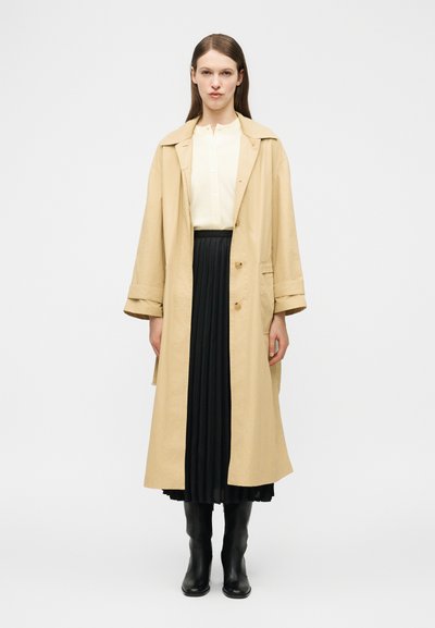 Femme portant un trench-coat beige sur un chemisier blanc, une jupe plissée noire et des bottes noires montant jusqu'aux genoux, debout devant un fond blanc uni.