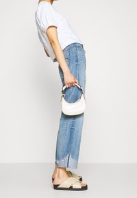 Sandalias de slide beige con una amplia tira cruzada sobre una suela negra, combinadas con jeans de un azul claro y una blusa blanca corta, sosteniendo un pequeño bolso blanco.