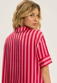 Oversized koszula z krótkim rękawem w naprzemiennie różowe i czerwone pionowe paski. Charakteryzuje się klasycznym kołnierzykiem i gładką, błyszczącą teksturą materiału.