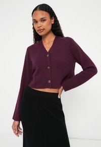 Kort kardigan i mörklila stickad tyg med ribbad nederkant och långa ärmar, med tre bruna knappar fram. Svart kjol.