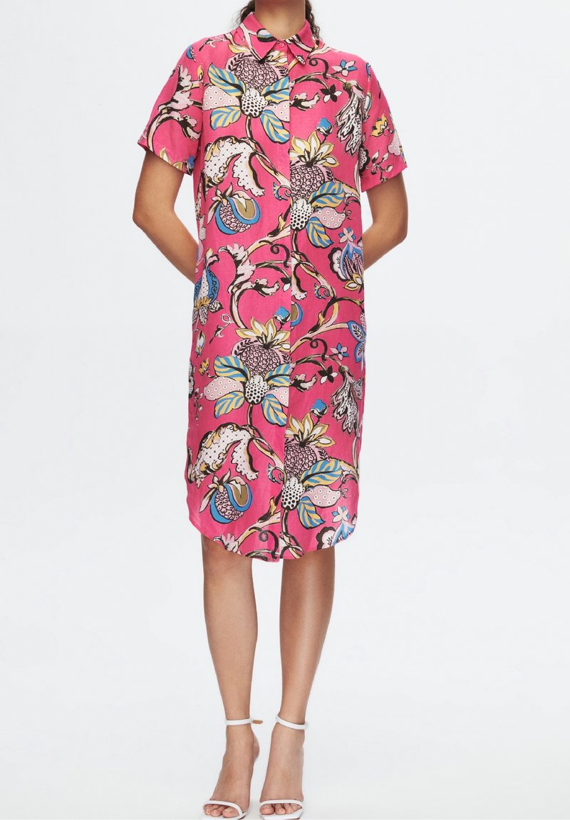 Femme portant une robe rose vif à manches courtes, longueur genoux, avec un motif floral et fruité coloré, associée à des sandales blanches à bride à la cheville.