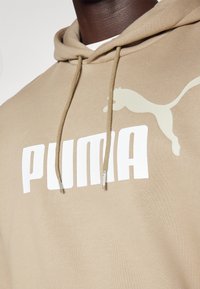 Beige Hoodie mit großem weißen "PUMA"-Logo und einer helleren beige Puma-Silhouette. Enthält Kordeln mit Plastikschnüren.
