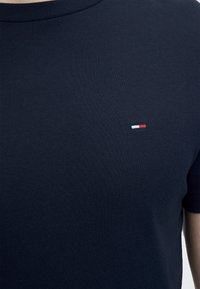 Κοντινό πλάνο ενός ατόμου που φοράει μπλε σκούρο μπλουζάκι με μικρό κόκκινο, λευκό και μπλε λογότυπο στην αριστερή πλευρά του στήθους.