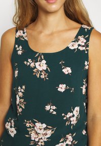 Femme portant un haut sans manches vert foncé avec des motifs floraux blancs et roses, mettant en valeur le cou et les épaules.