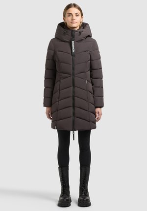 Mujer con un abrigo largo acolchado marrón oscuro con capucha, leggings negros y botas negras con cordones, de pie sobre un fondo sencillo.