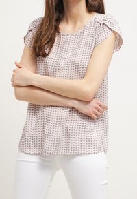 Blusa rosa claro con pequeño estampado floral, mangas cortas con volantes y un corte relajado. La tela parece suave y ligera. Se ven jeans blancos.
