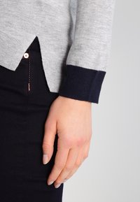 Jersey gris de punto con puño acanalado azul marino y abertura lateral, combinado con pantalones oscuros. Primer plano de una mano apoyada sobre la prenda.