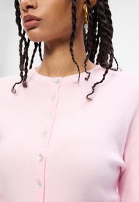 Ανοιχτό ροζ πλεκτό ζακέτα με στρογγυλή λαιμόκοψη, διαθέτοντας μια σειρά λευκών κουμπιών κατά μήκος του μπροστινού μέρους και ριμπ πλέξη στις άκρες.